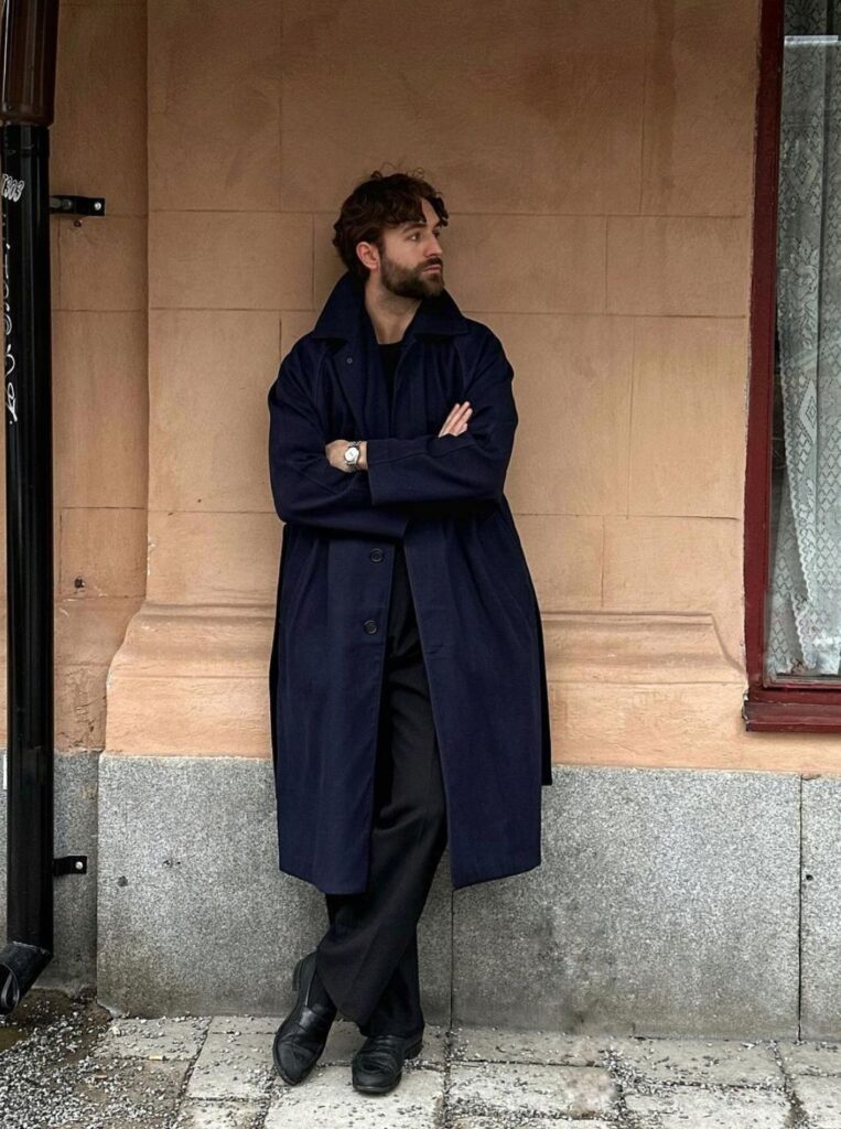 Man wearing a navy overcoat, black t-shirt and black casual trousers.